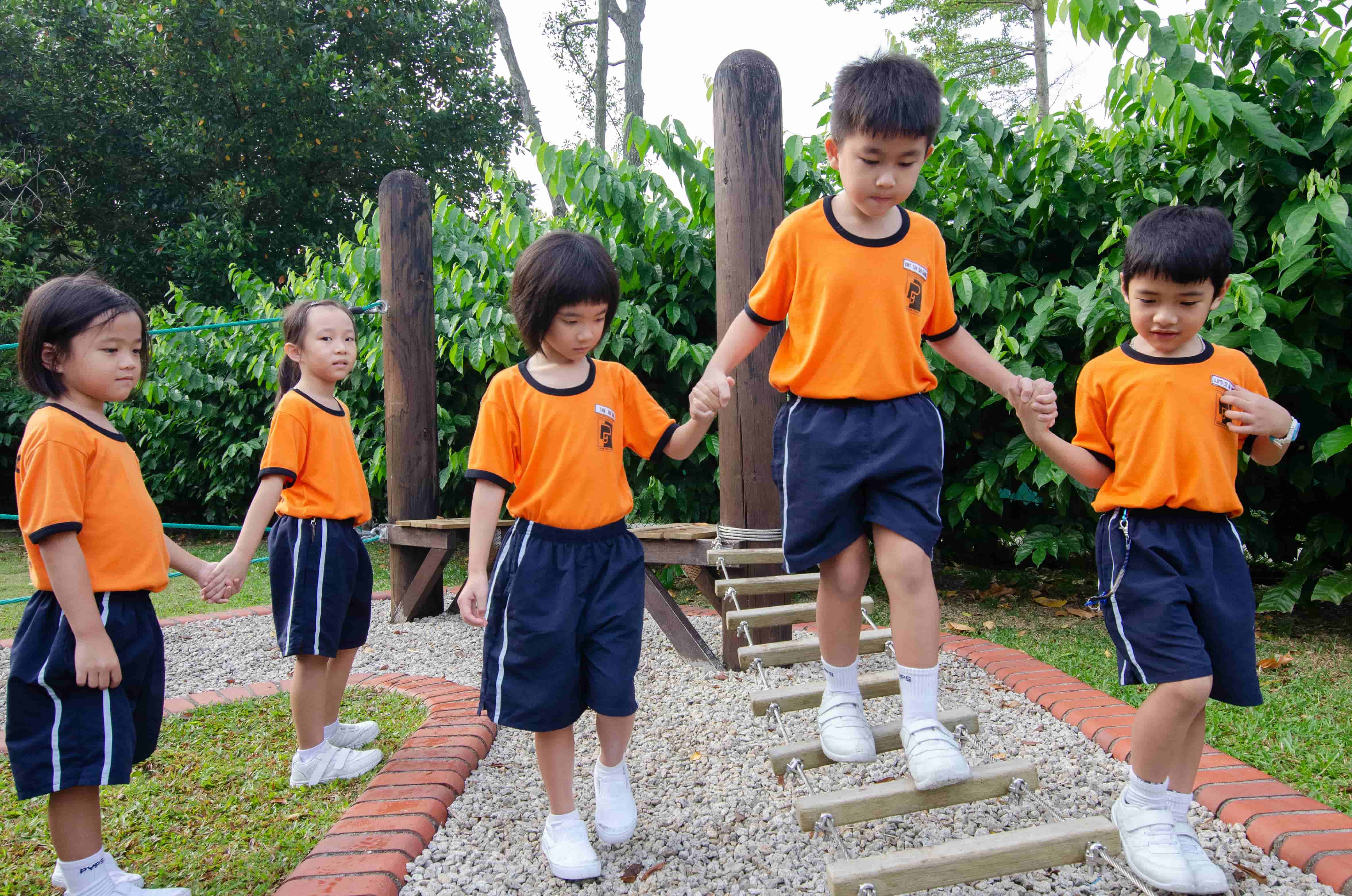 Five young children in matching orange shirts and navy shorts hold hands while navigating a wooden rope ladder outdoors.